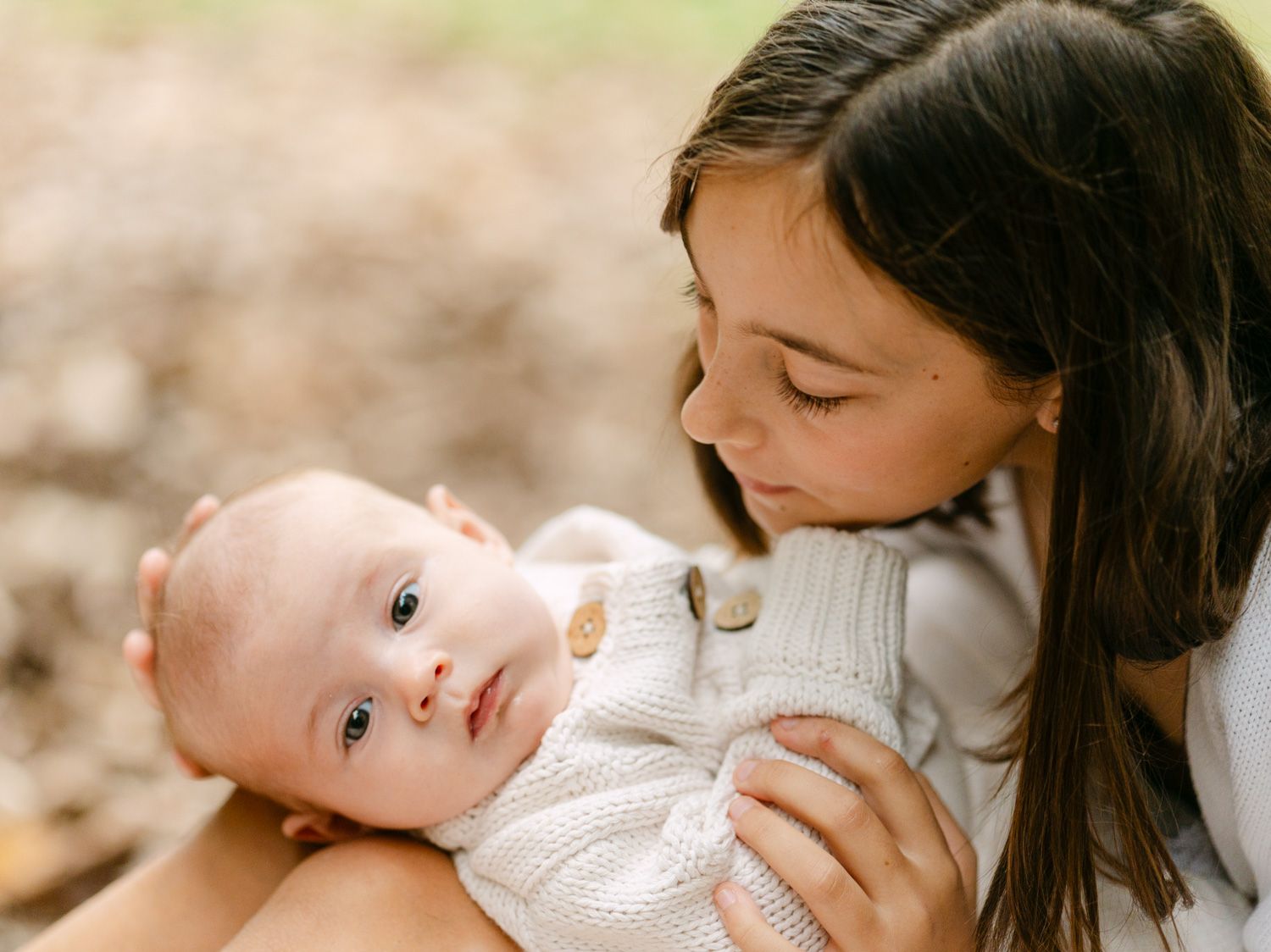 A young woman tenderly holds and gazes at her newborn baby outdoors in a warm, bokeh nature setting.