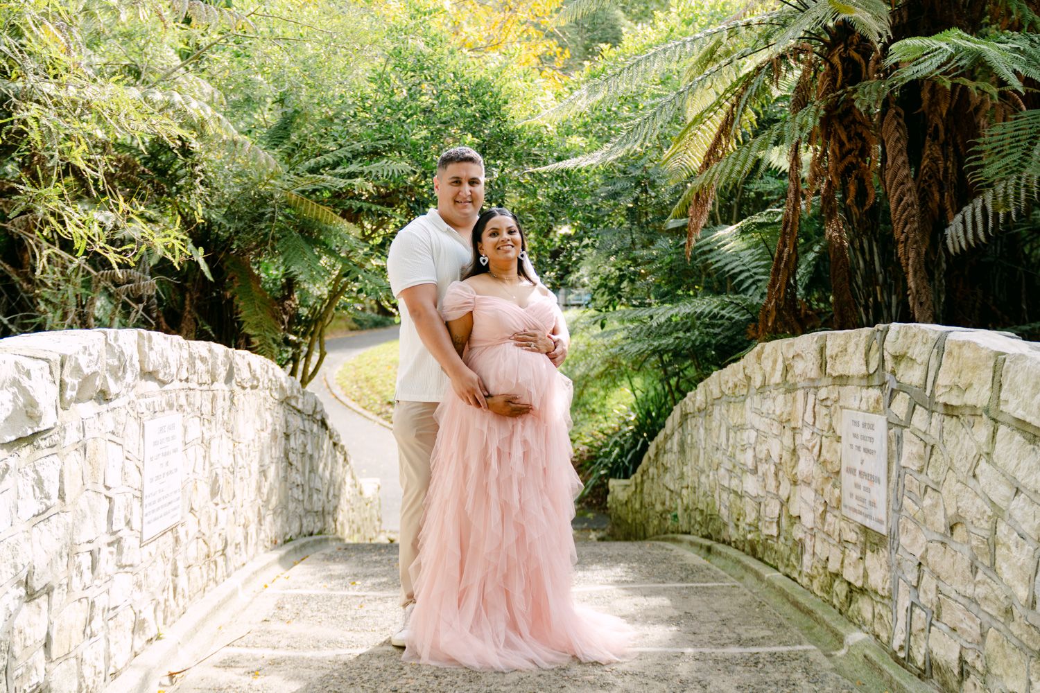 Expectant couple smiles on stone bridge surrounded by tropical greenery, woman in pink gown.