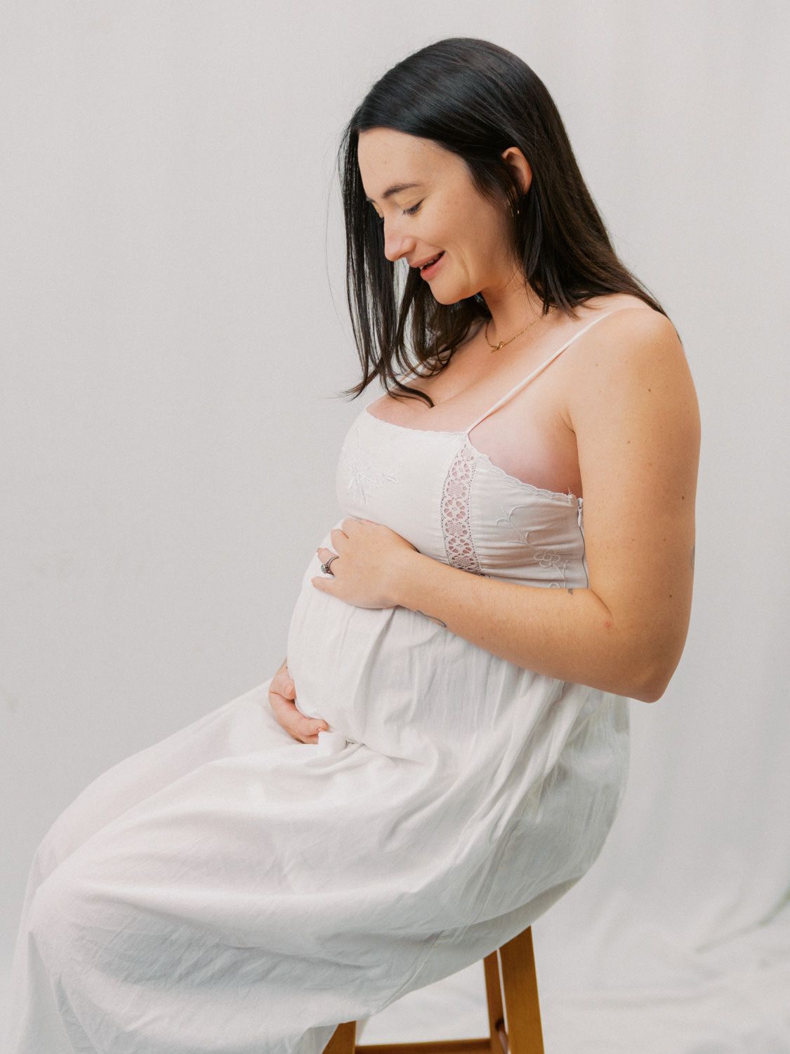 Pregnant woman in white dress sitting on stool, cradling baby bump, smiling softly in a bright studio setting.