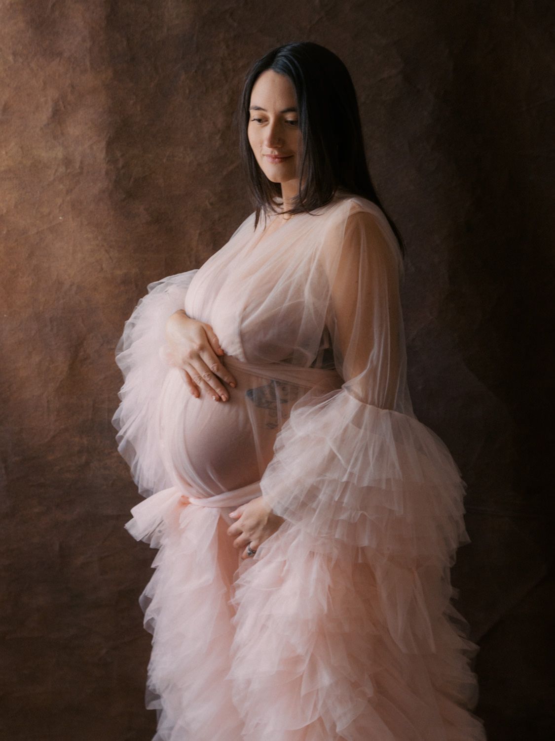 Pregnant woman in sheer pink ruffle maternity gown cradling baby bump in professional studio portrait.