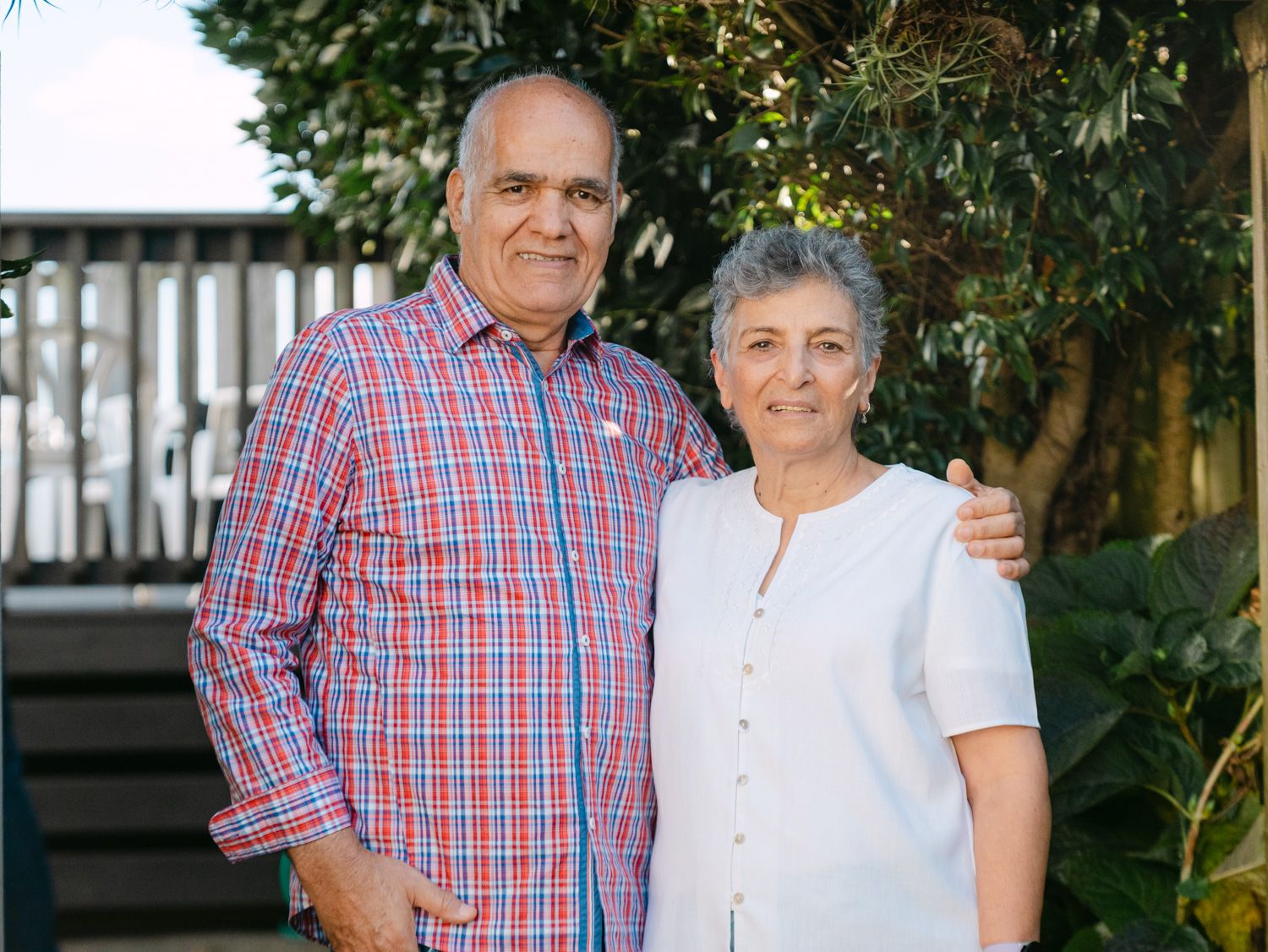 Smiling older couple posing outdoors together, man in plaid shirt with arm around woman in white top.