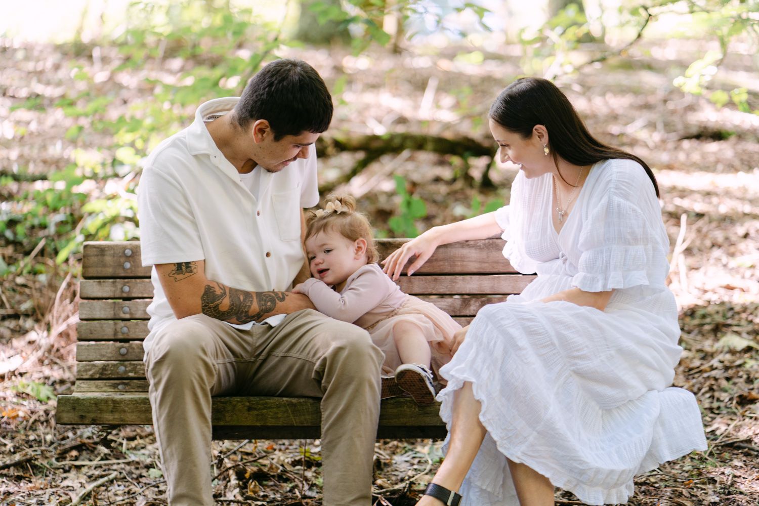 Family of three sitting on a wooden bench in a forest, parents lovingly attending to their toddler child.