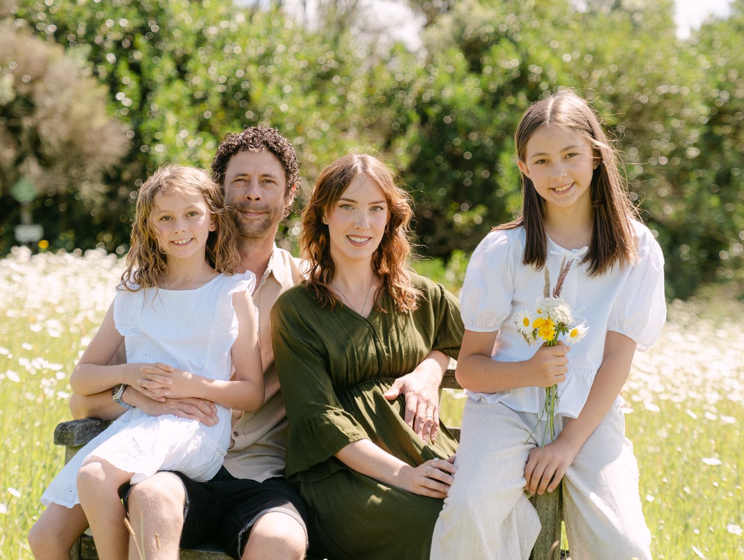 Happy family of four posing outdoors in a sunny wildflower meadow, smiling together on a wooden bench.