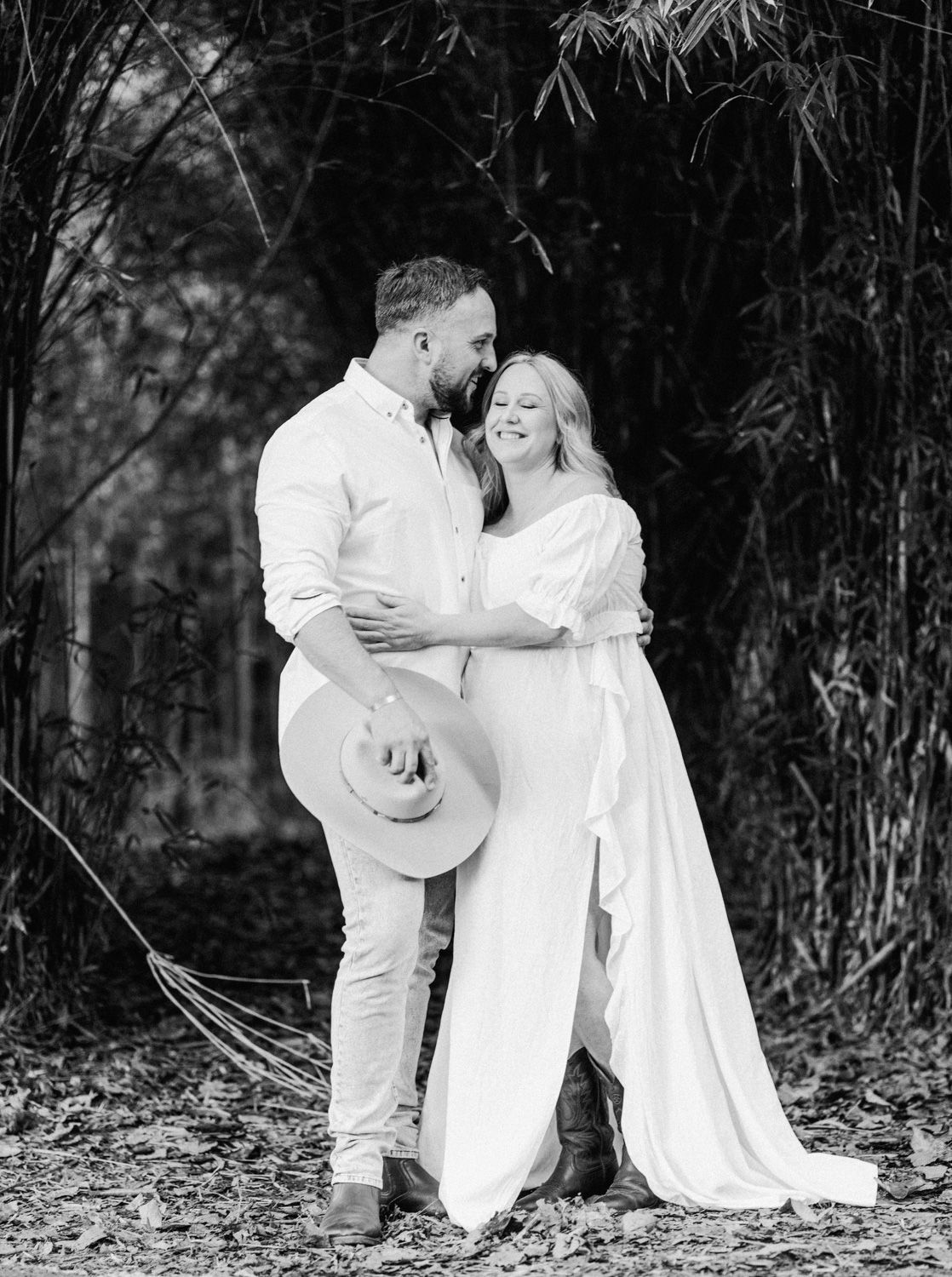 Black and white photo of a couple in white outfits embracing outdoors among bamboo trees.