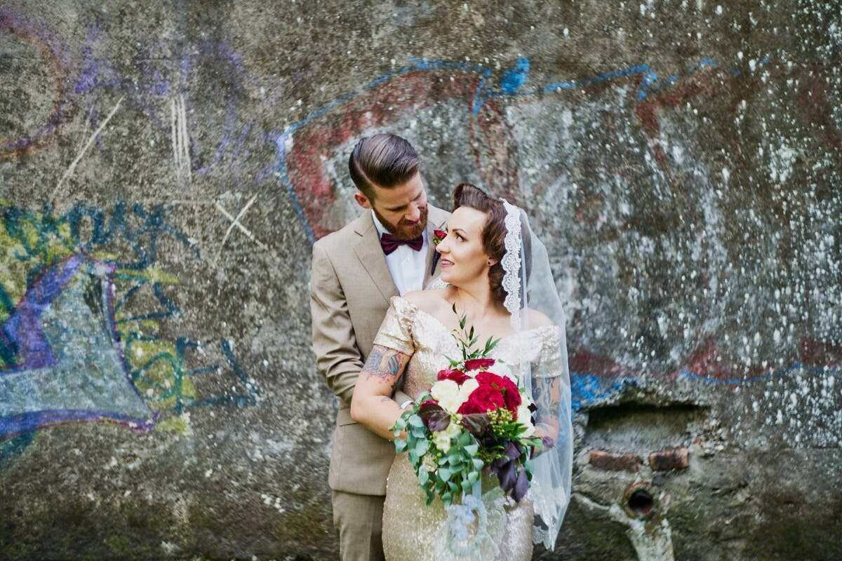 Bride and groom embrace before graffiti wall, bride holding red and white bouquet wearing lace gown and veil.
