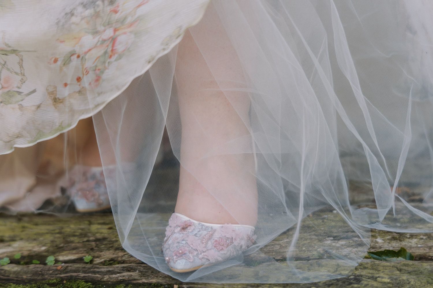 Close-up of a bride wearing pink floral heels and a flowing white tulle wedding gown standing on rustic stone.