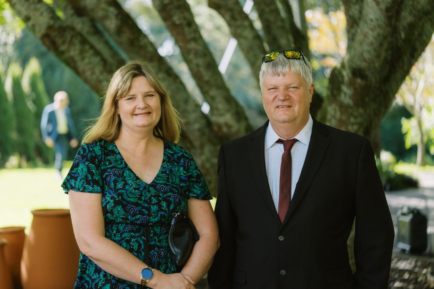 Couple smiling outdoors near a large tree, woman in floral dress and man in black suit with red tie.