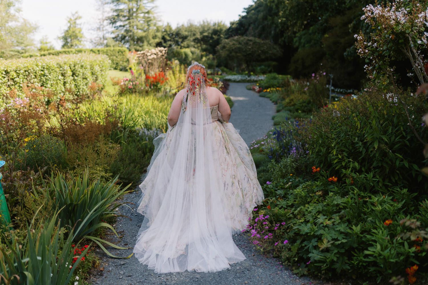 Bride in flowing white tulle gown walks through lush garden path surrounded by colorful flowers on wedding day.
