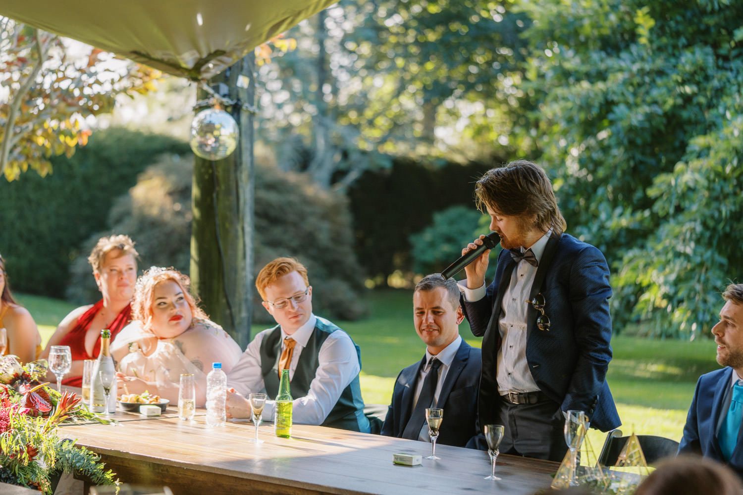 Wedding speech delivered outdoors under a tent, with guests smiling at a decorated reception table in a garden setting.