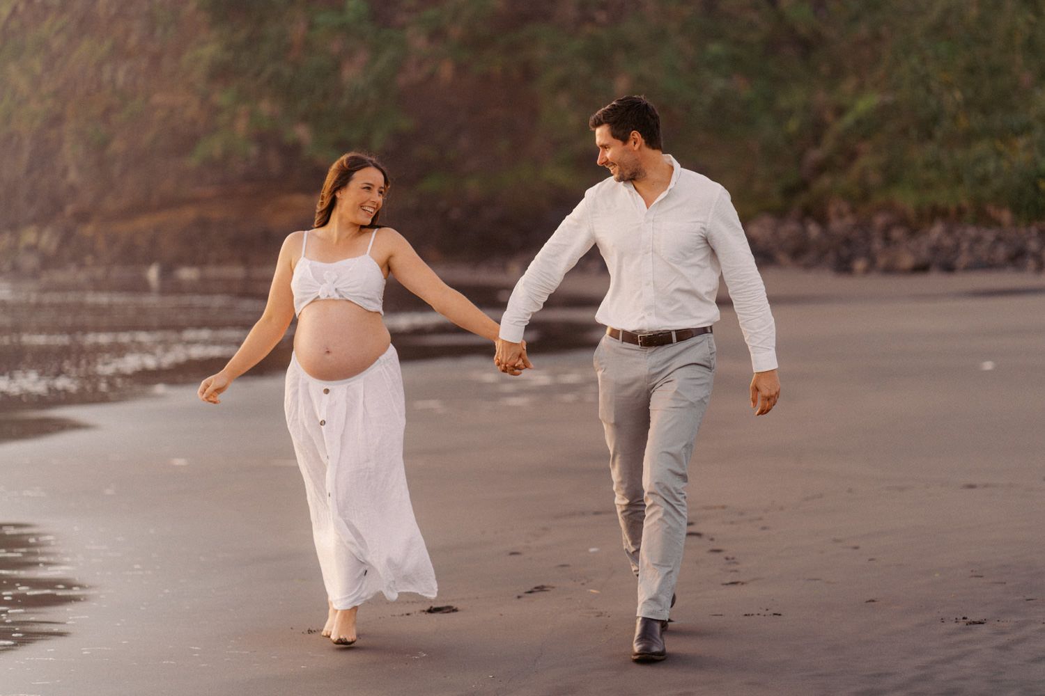 Pregnant couple holding hands walking on a beach, woman in white dress, man in white shirt and gray pants.