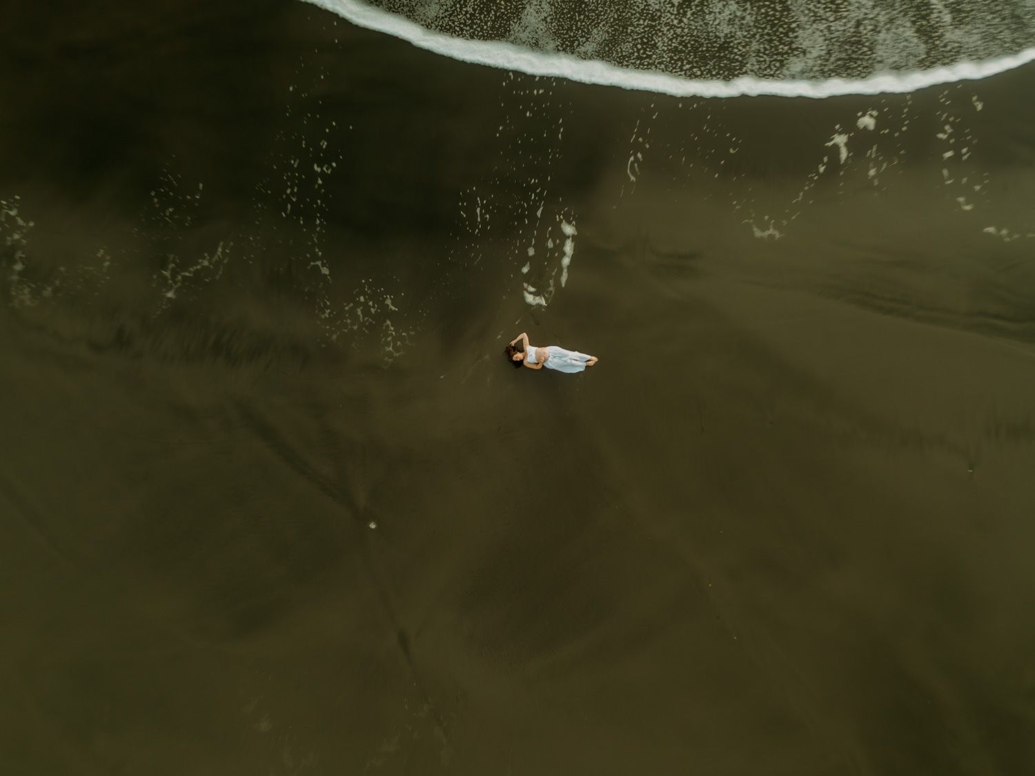 Aerial drone view of a person in a white dress lying on a dark sandy beach near ocean waves.