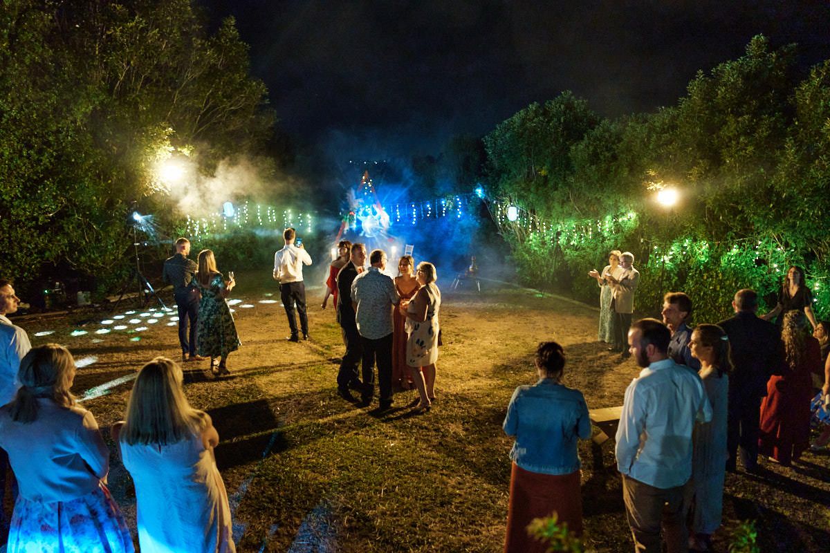 Outdoor evening wedding reception with guests gathered under glowing lights surrounded by lush greenery and colorful fog effects.