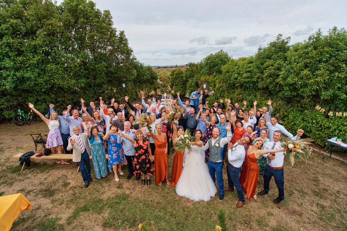 Large wedding group photo outdoors in a lush garden, bride and groom surrounded by cheering guests.
