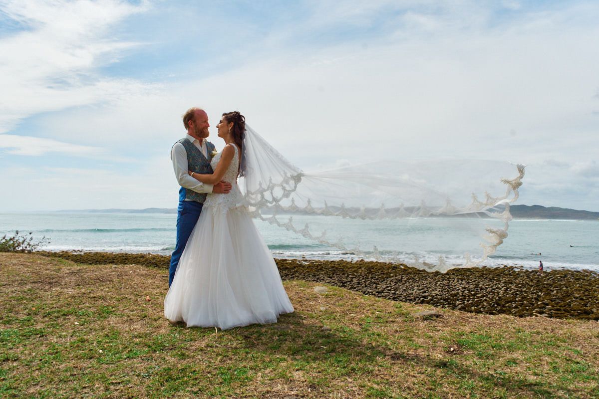 Bride and groom embrace by the ocean, her veil flowing in the wind, on a sunny wedding day near the coast.
