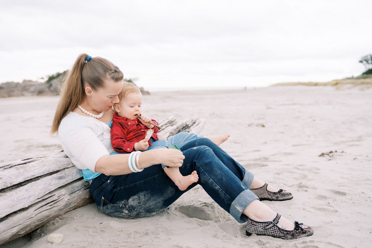A mother sits on driftwood at the beach, lovingly holding her baby in a red outfit on a cloudy day.