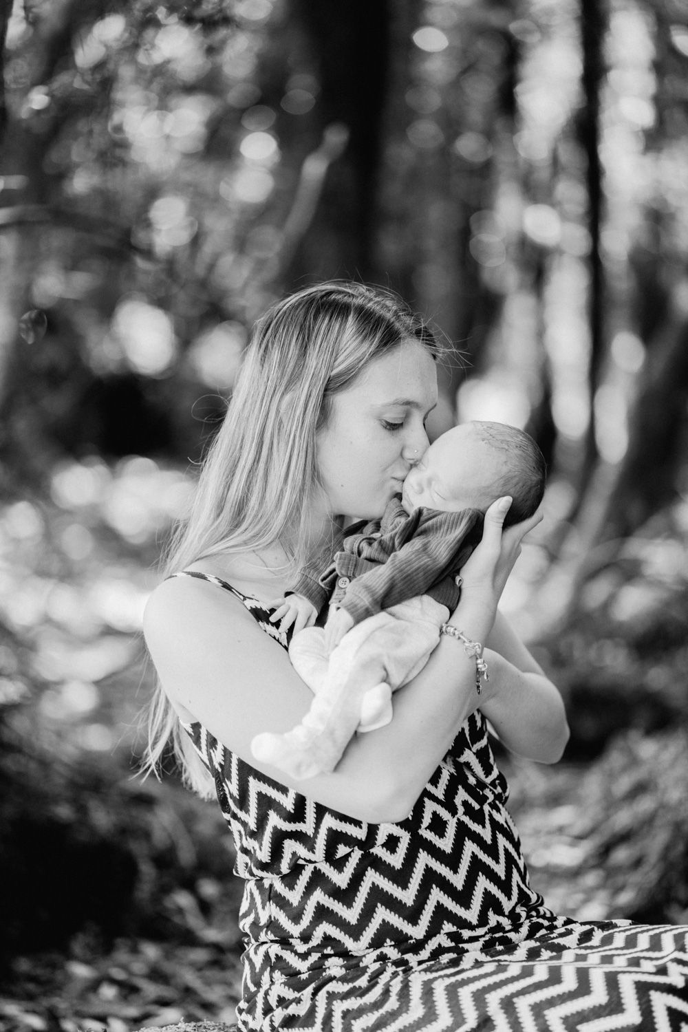 Black and white photo of a mother kissing her newborn baby outdoors in a lush forest setting.