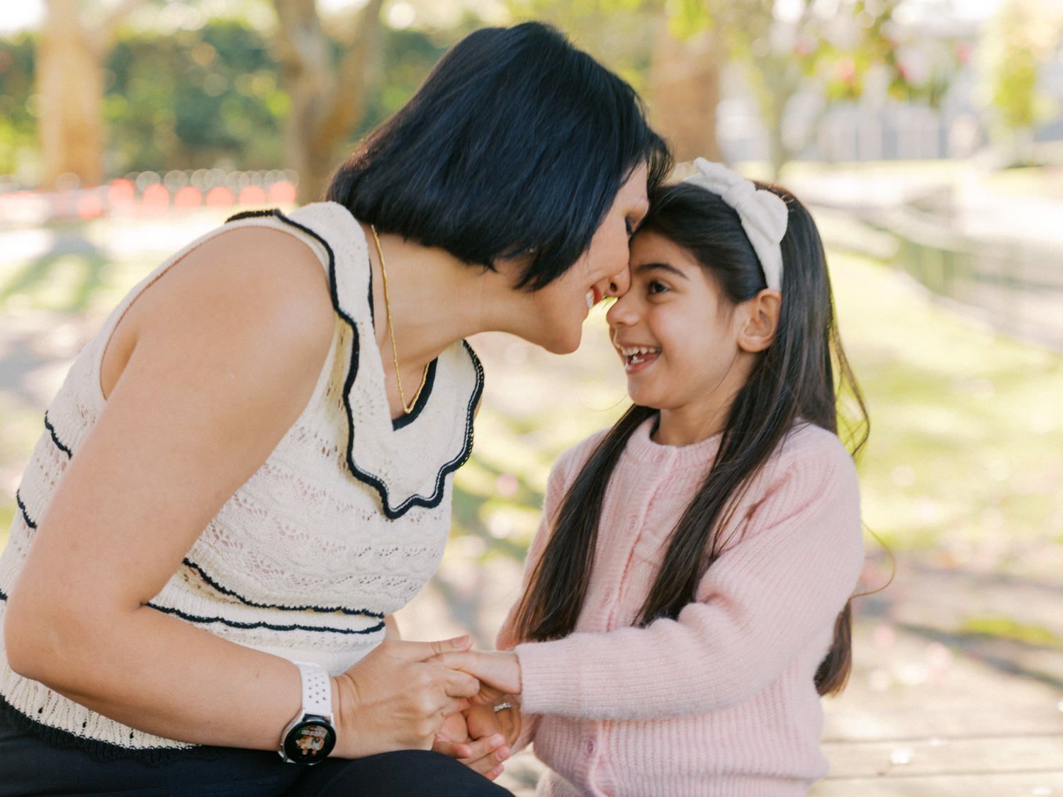 A mother and daughter share a tender nose-to-nose moment while holding hands outdoors in a sunny park.