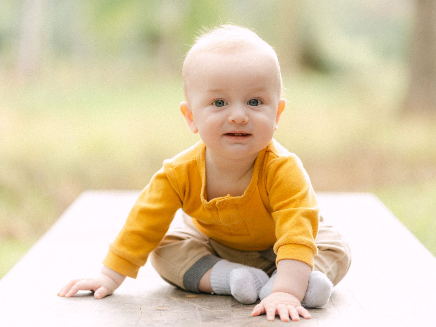 Adorable baby in a yellow outfit sitting outdoors on a surface, smiling at the camera with a green background.