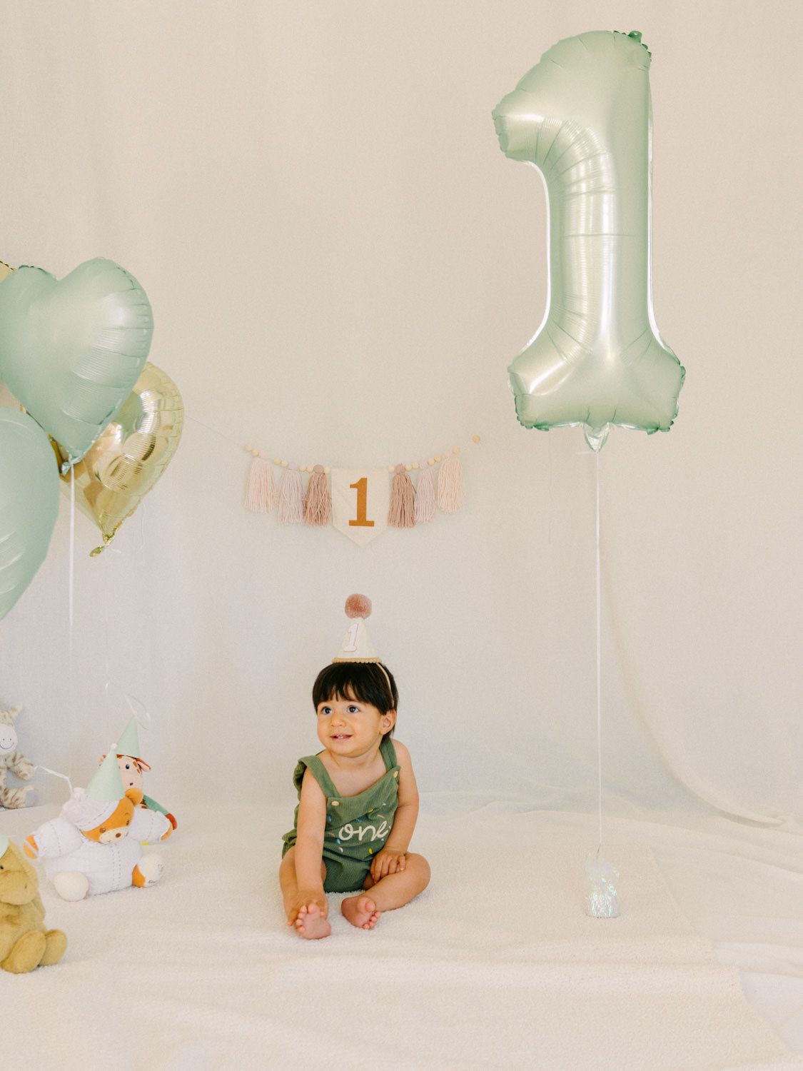 Baby girl sits on white floor near number 1 balloon and tassel banner at her first birthday party.