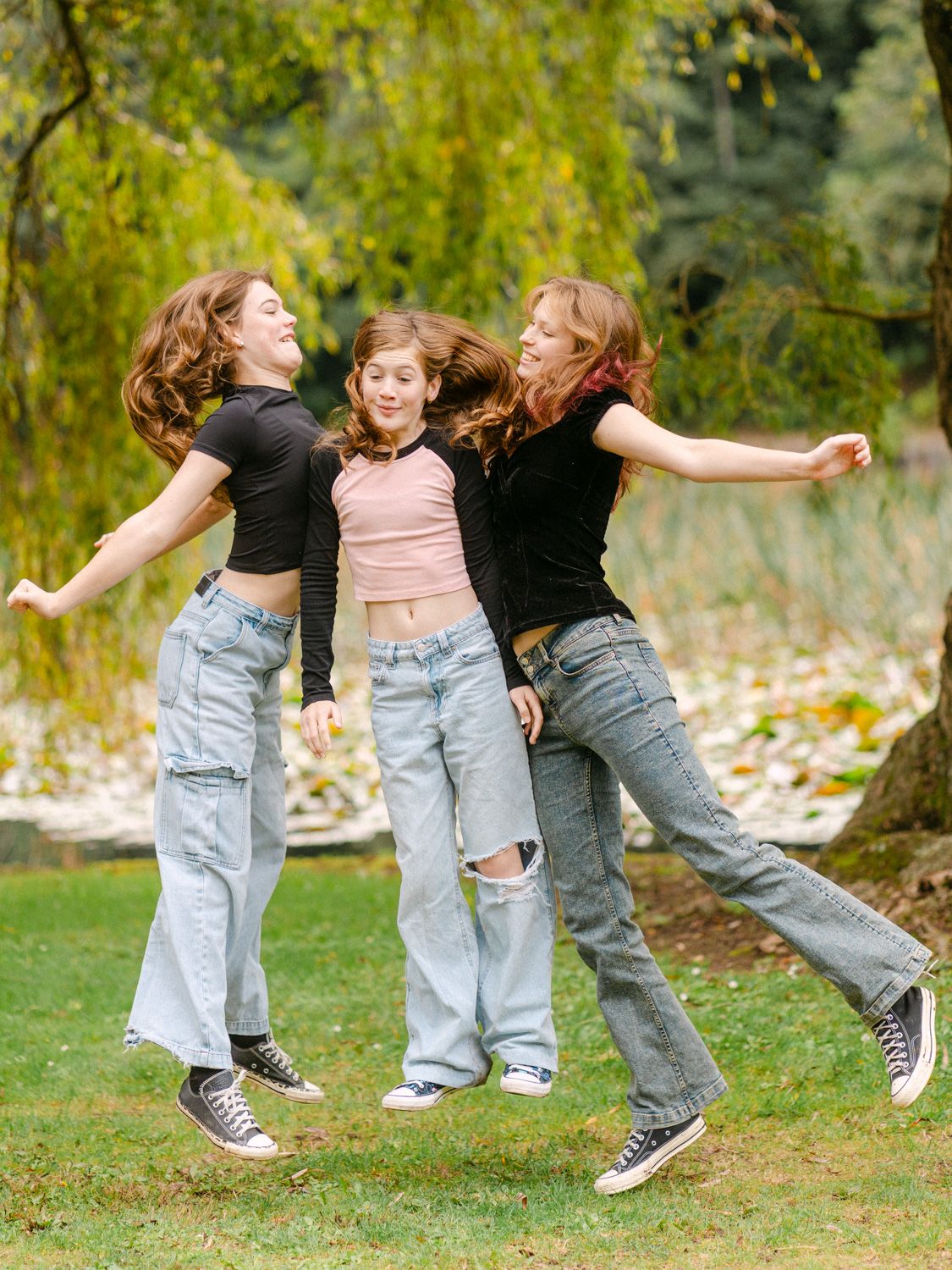 Three teenage girls in crop tops and wide-leg jeans jump together outdoors in a lush green park setting.