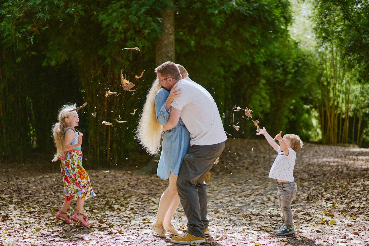 Family dancing outdoors while children toss autumn leaves on a tree-lined path.