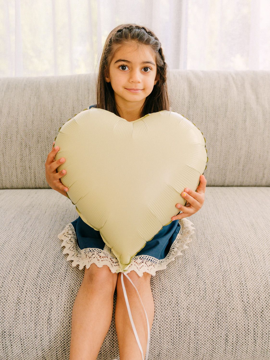 Young girl sitting on a gray sofa holding a large cream heart-shaped balloon, wearing a blue dress with lace trim.