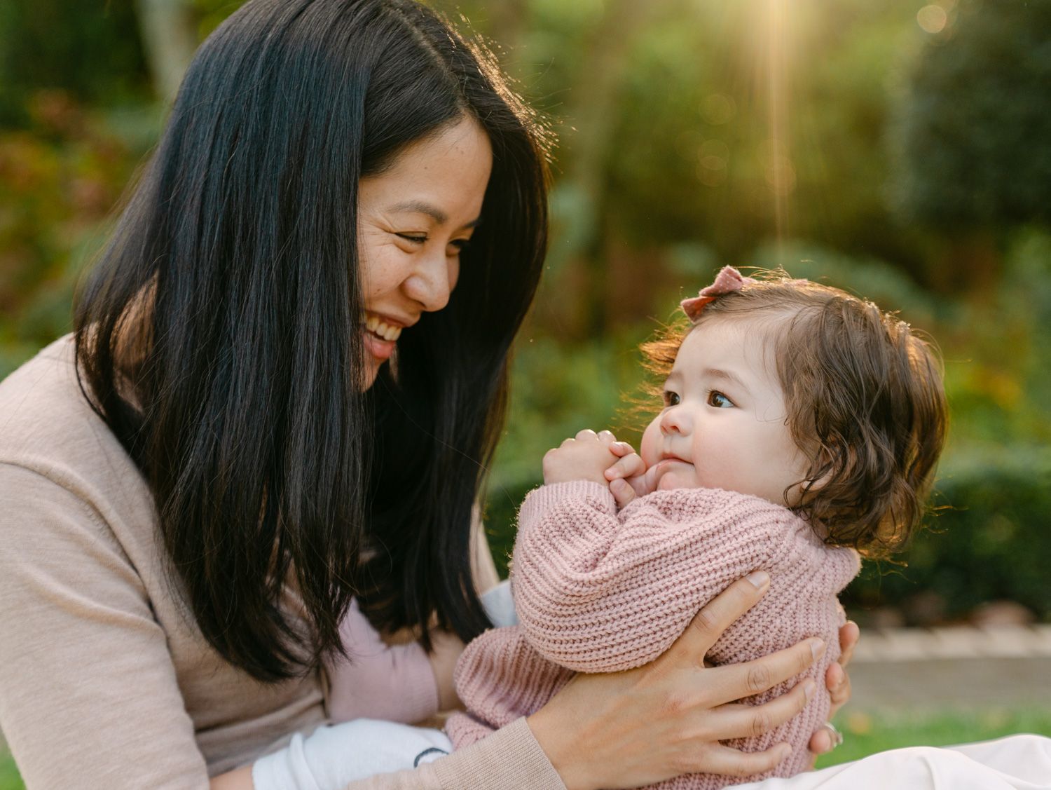 A smiling Asian mother tenderly holds her adorable baby girl in a pink sweater outdoors in a sunlit garden.