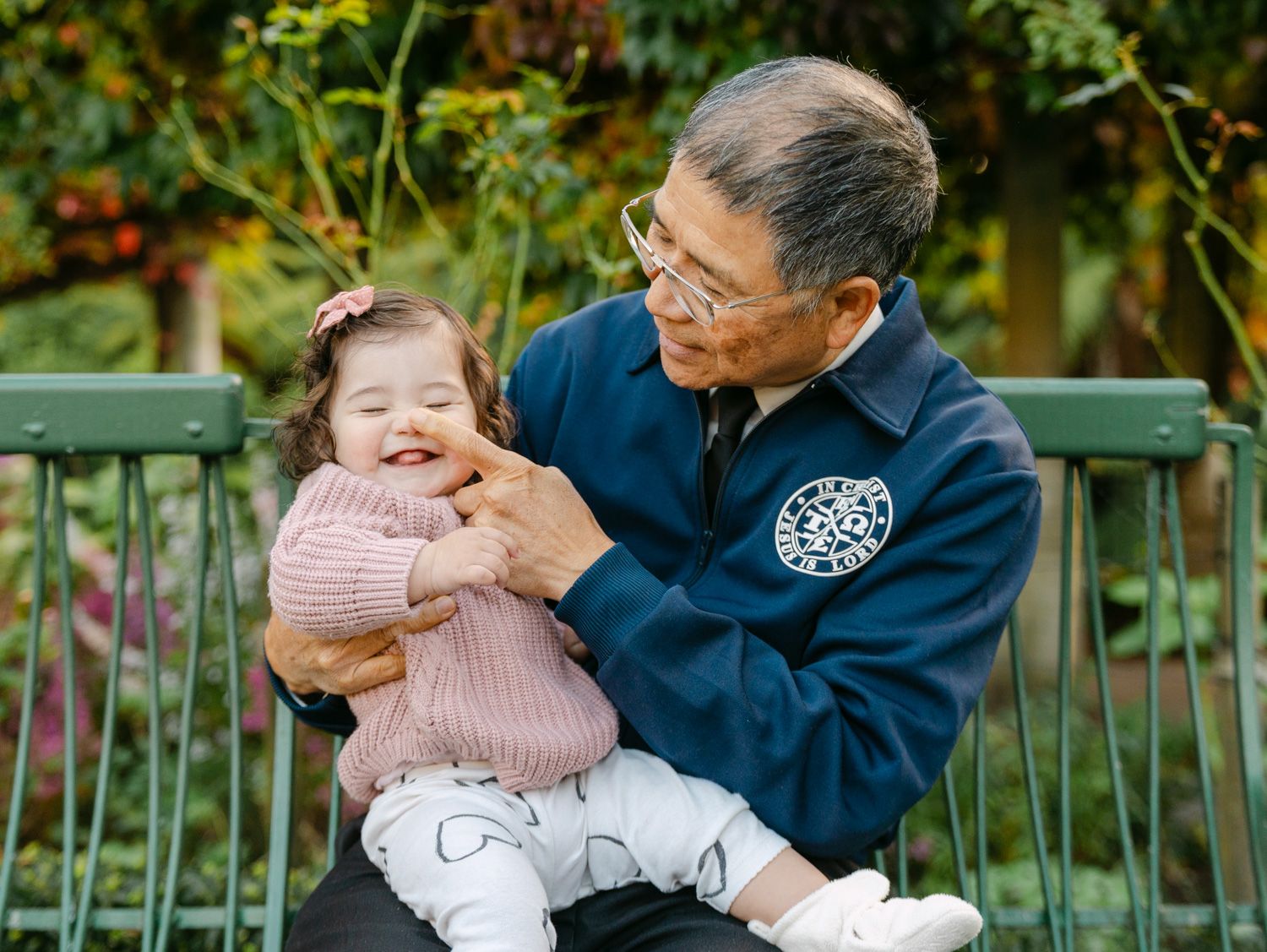 Elderly grandfather joyfully holding a laughing baby girl on a park bench surrounded by lush greenery.