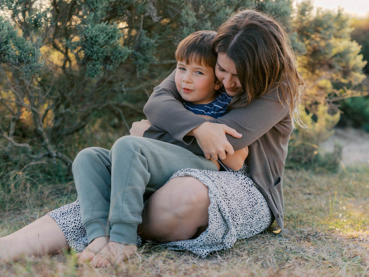 Mother hugging her young son while sitting on grass surrounded by green trees outdoors.