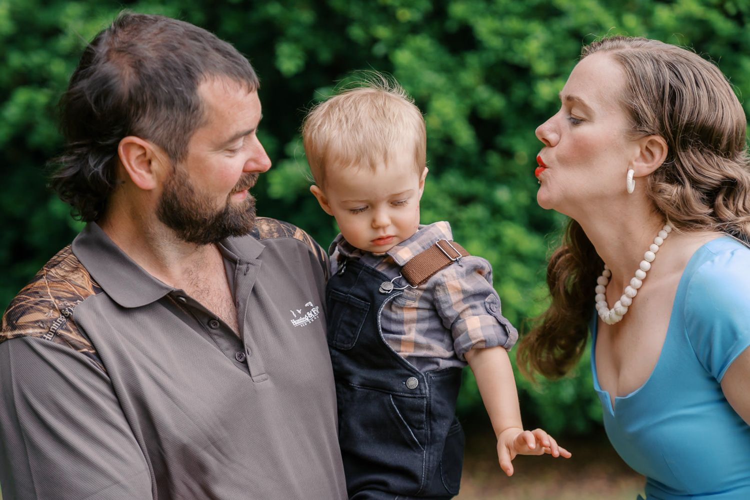 A man and woman with pearl necklace blowing a kiss to a toddler boy outdoors with green foliage background.