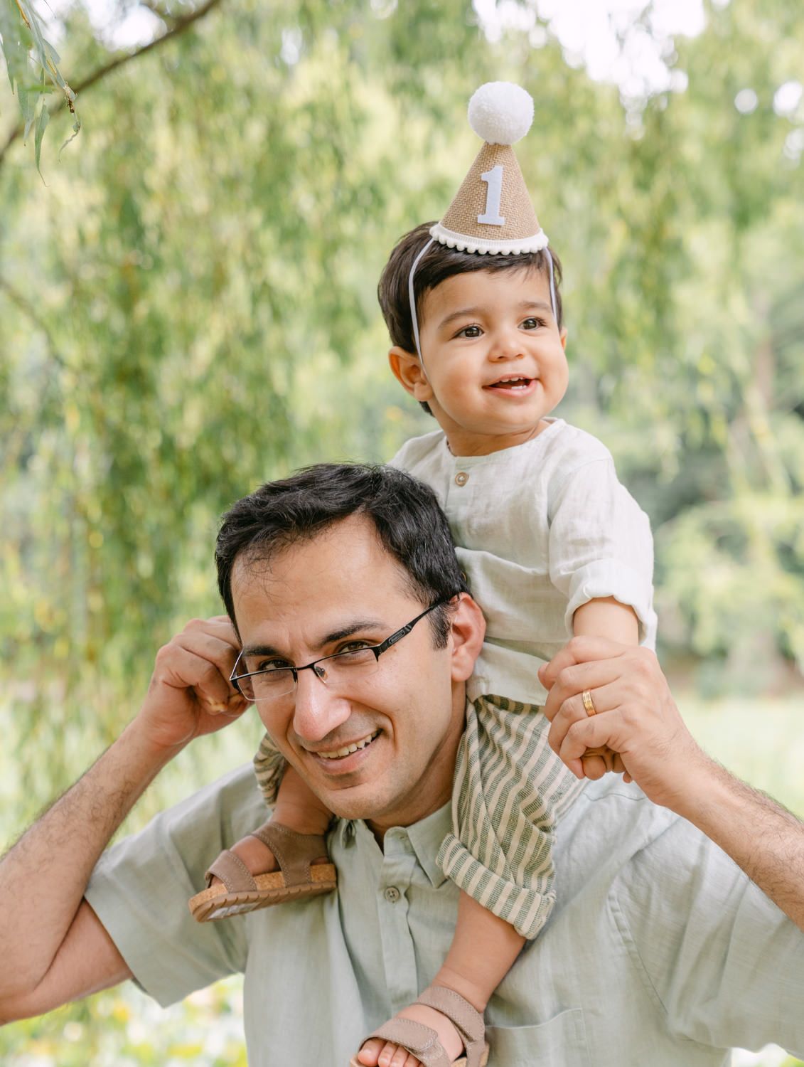 A smiling man carries a happy toddler wearing a number one birthday hat on his shoulders outdoors.