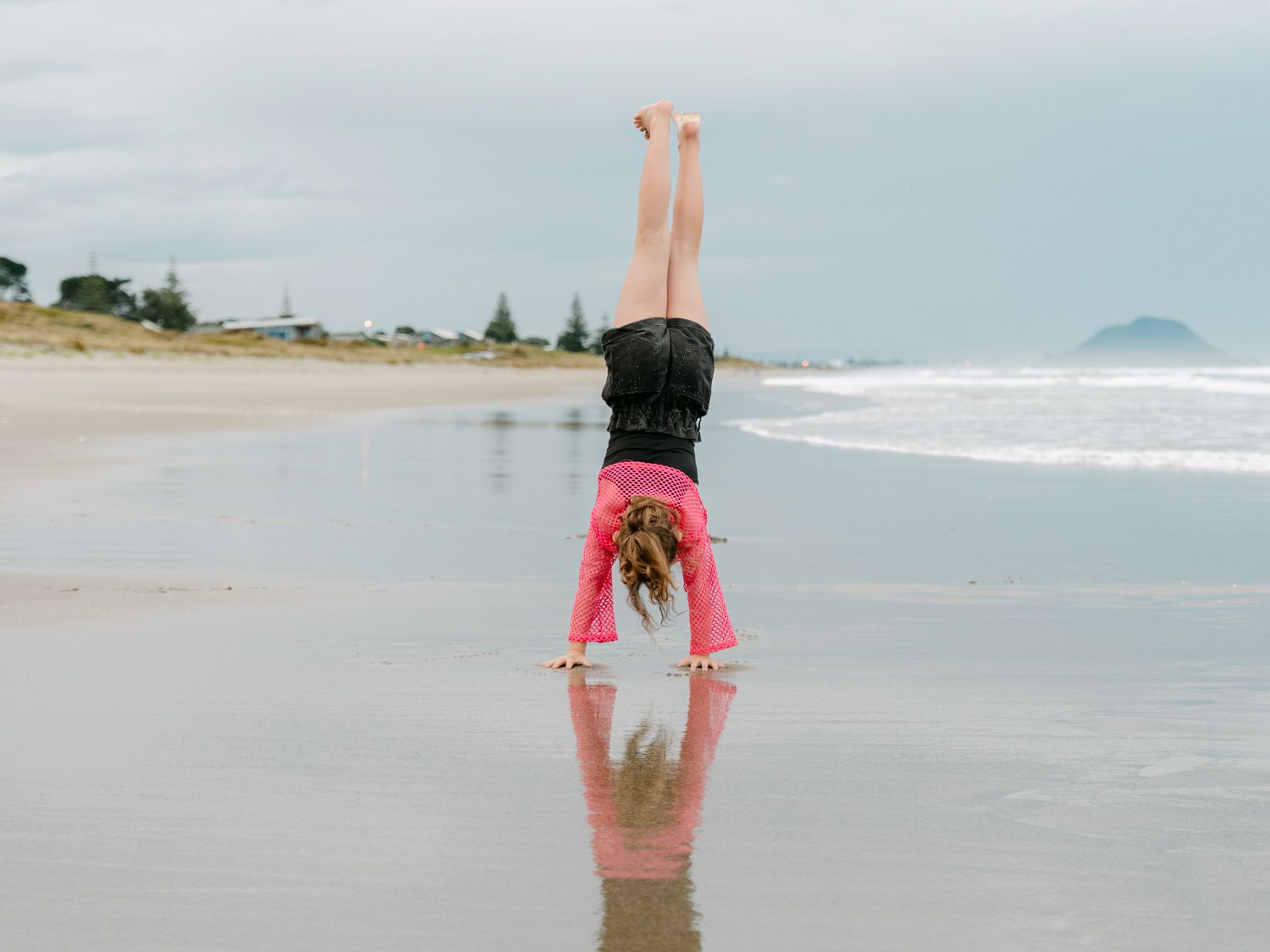 Girl doing a handstand on a reflective wet beach with ocean waves and cloudy sky in the background.