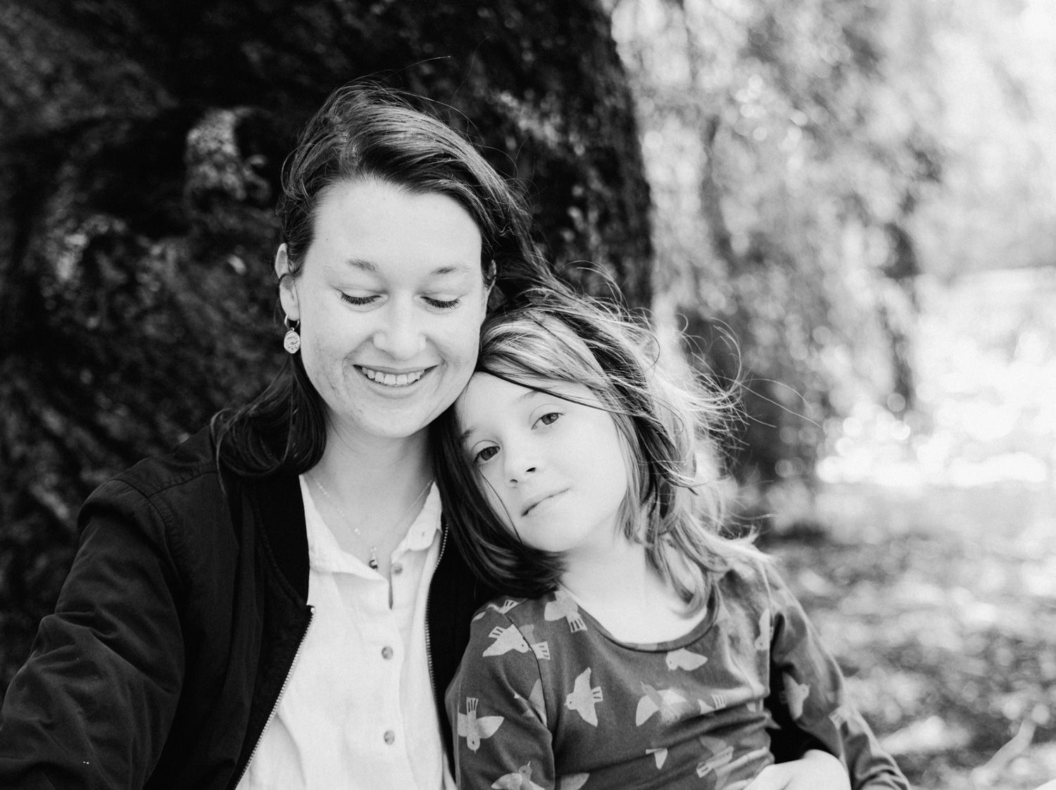Black and white photo of a smiling mother and young child leaning together outdoors near a tree.