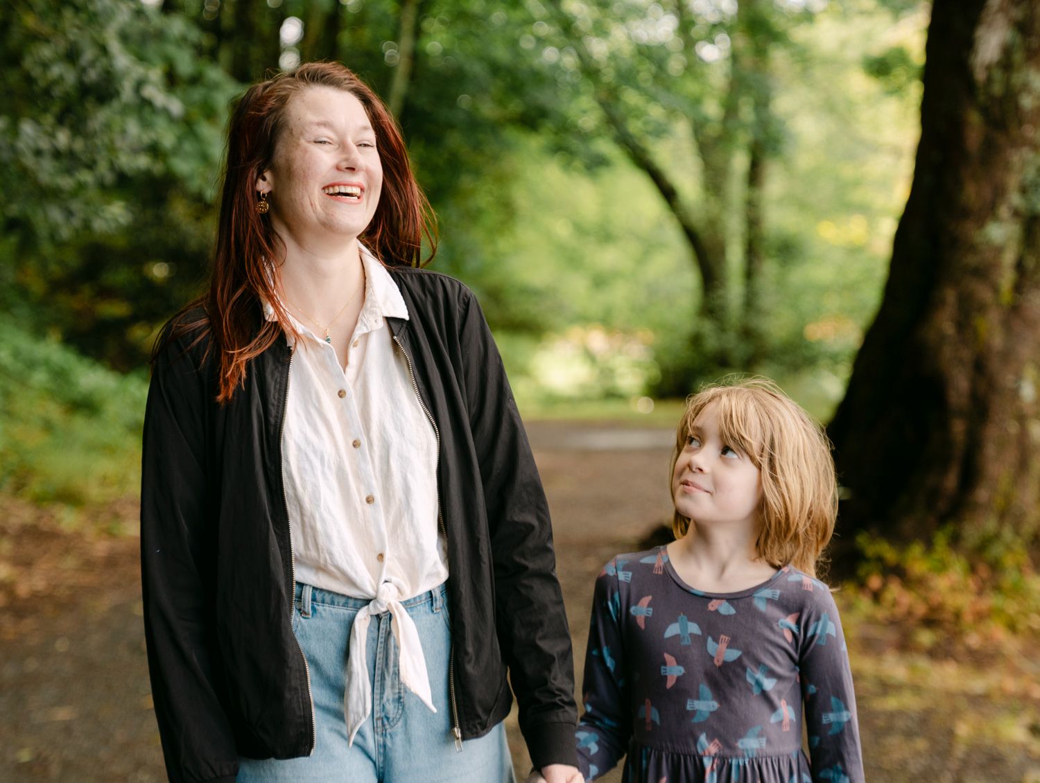 A woman and young girl walk hand in hand on a forest path, smiling together outdoors.