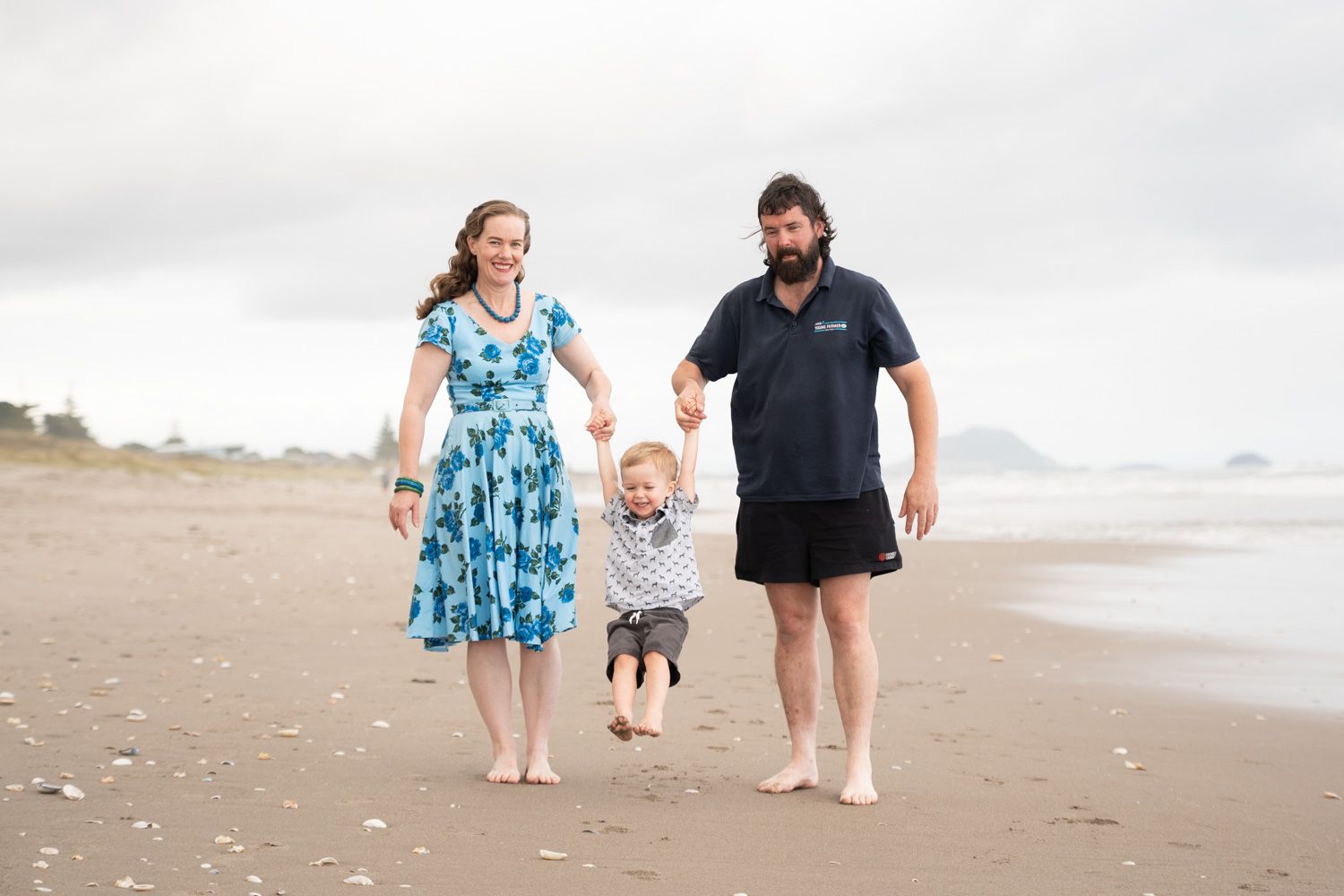 Family of three walking on a sandy beach, parents swinging toddler between them on a cloudy day.