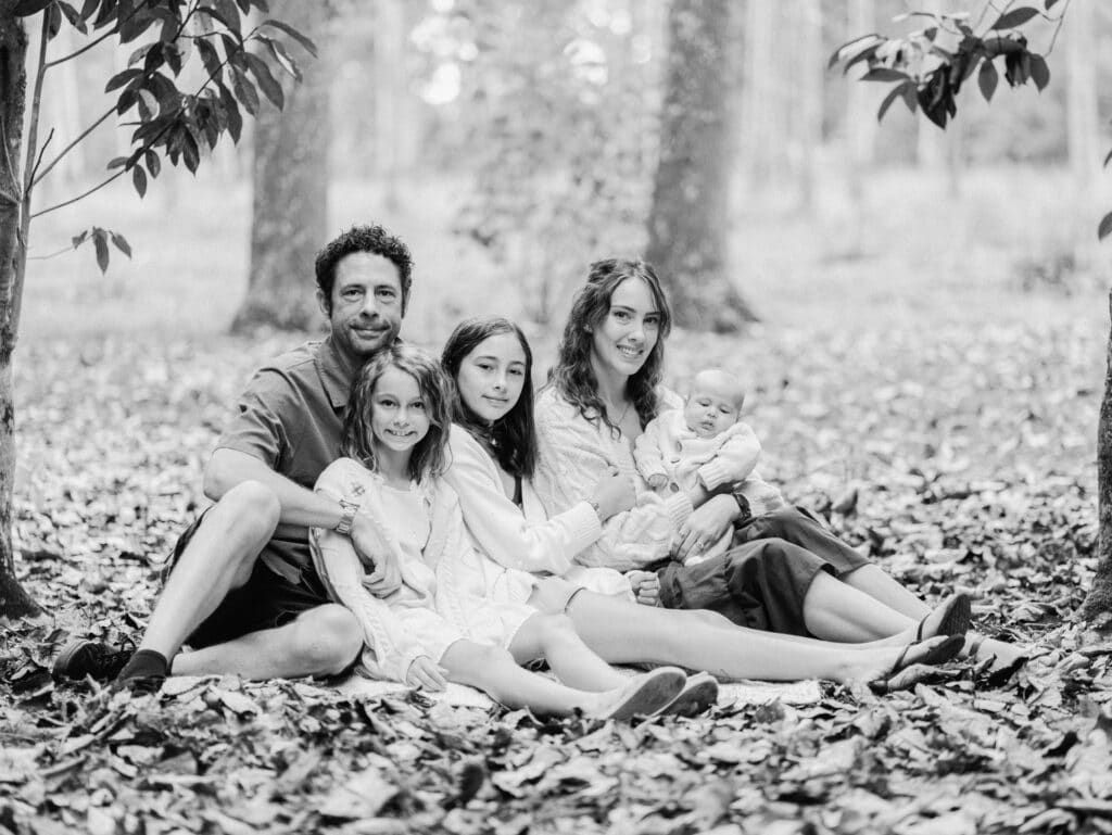 Black and white family portrait of parents with three children sitting on fallen leaves in a forest.
