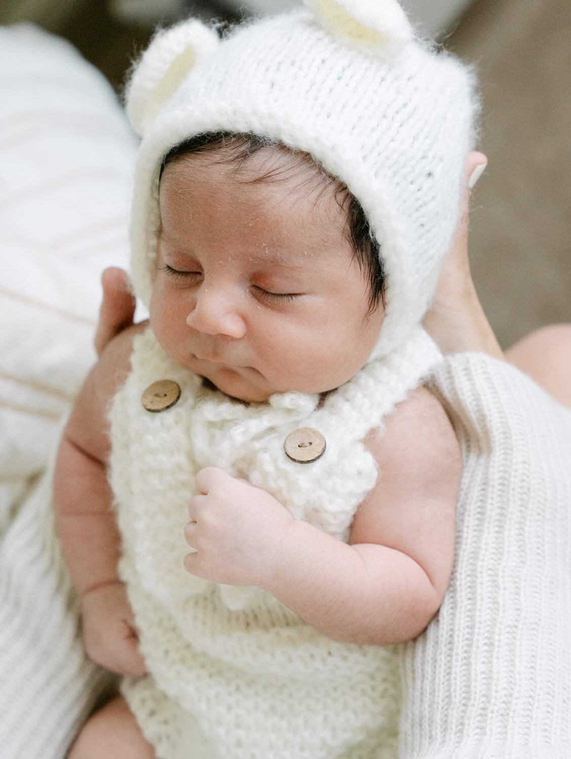Sleeping newborn baby wearing a white knit bear bonnet and cream button romper, cradled in loving arms.