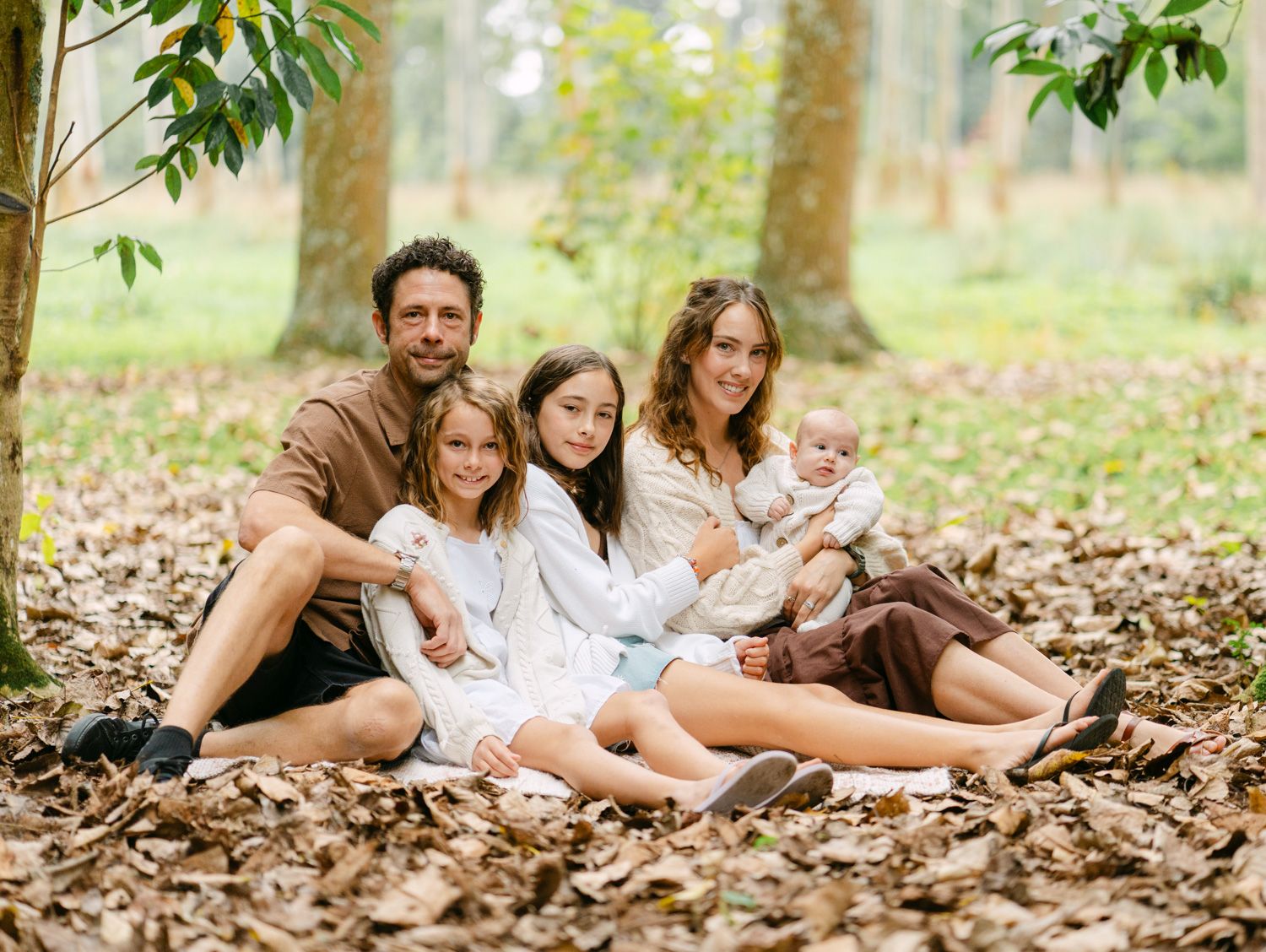 Family of four sitting on autumn leaves in a forest, parents with two daughters smiling outdoors.