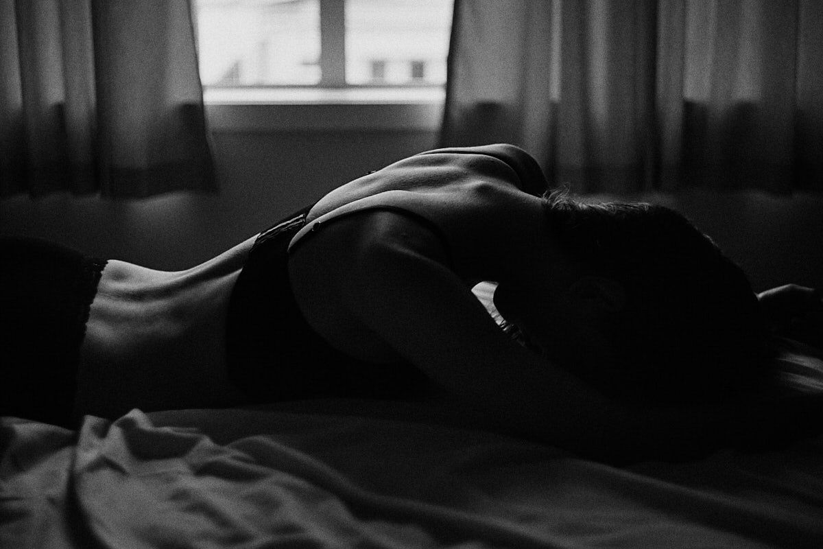Black and white photo of a person lying on a bed near curtained window in artistic boudoir style.