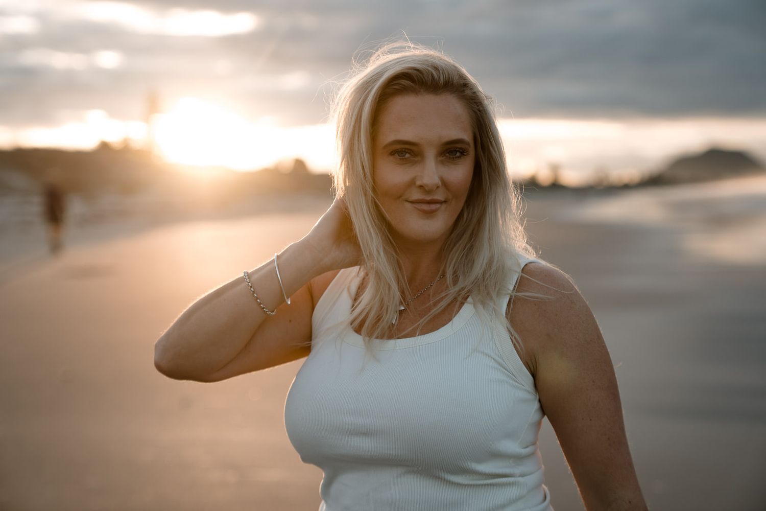 Blonde woman in white tank top posing on beach at golden sunset hour.