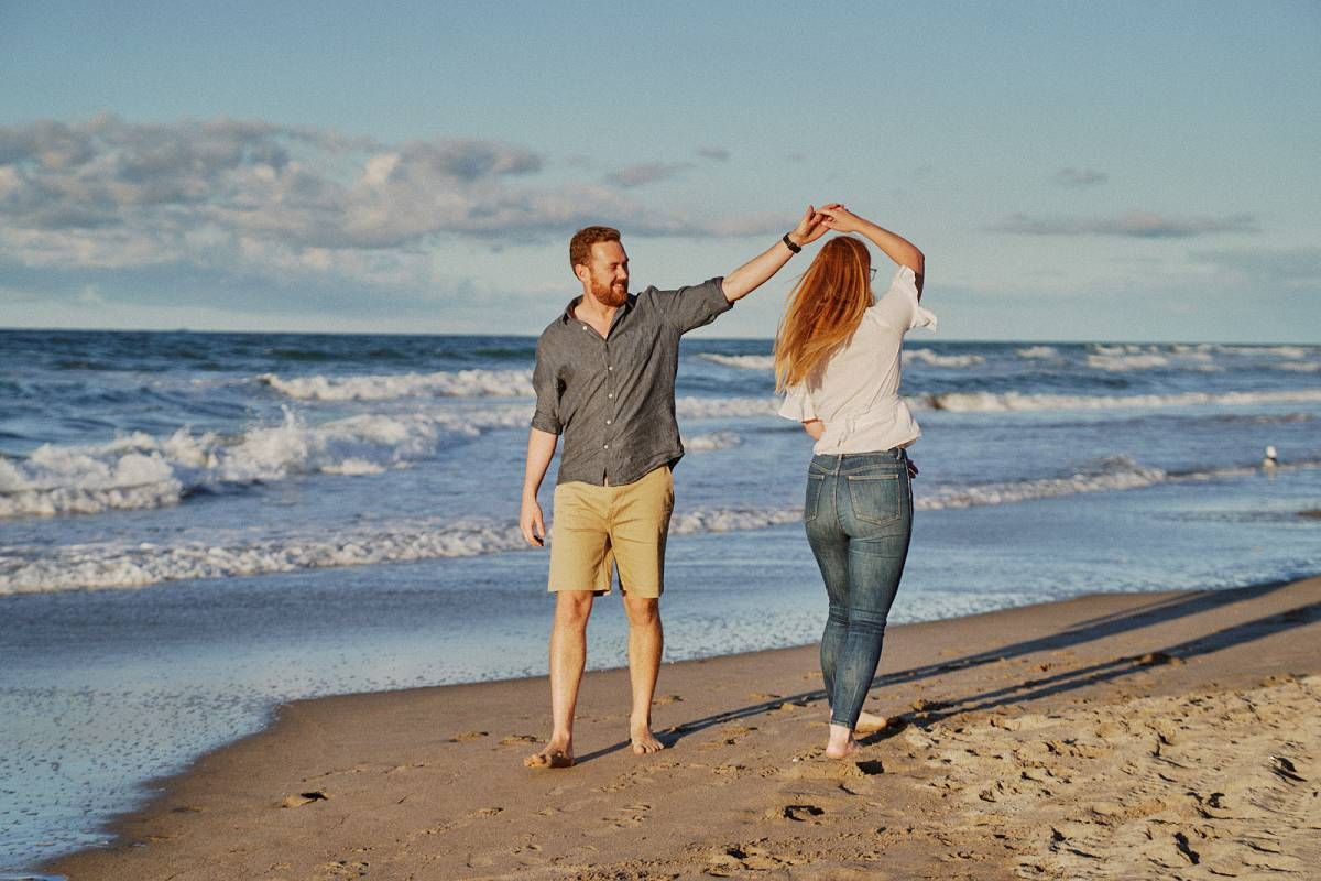 Couple dancing barefoot on a sandy beach at sunset with ocean waves in the background.