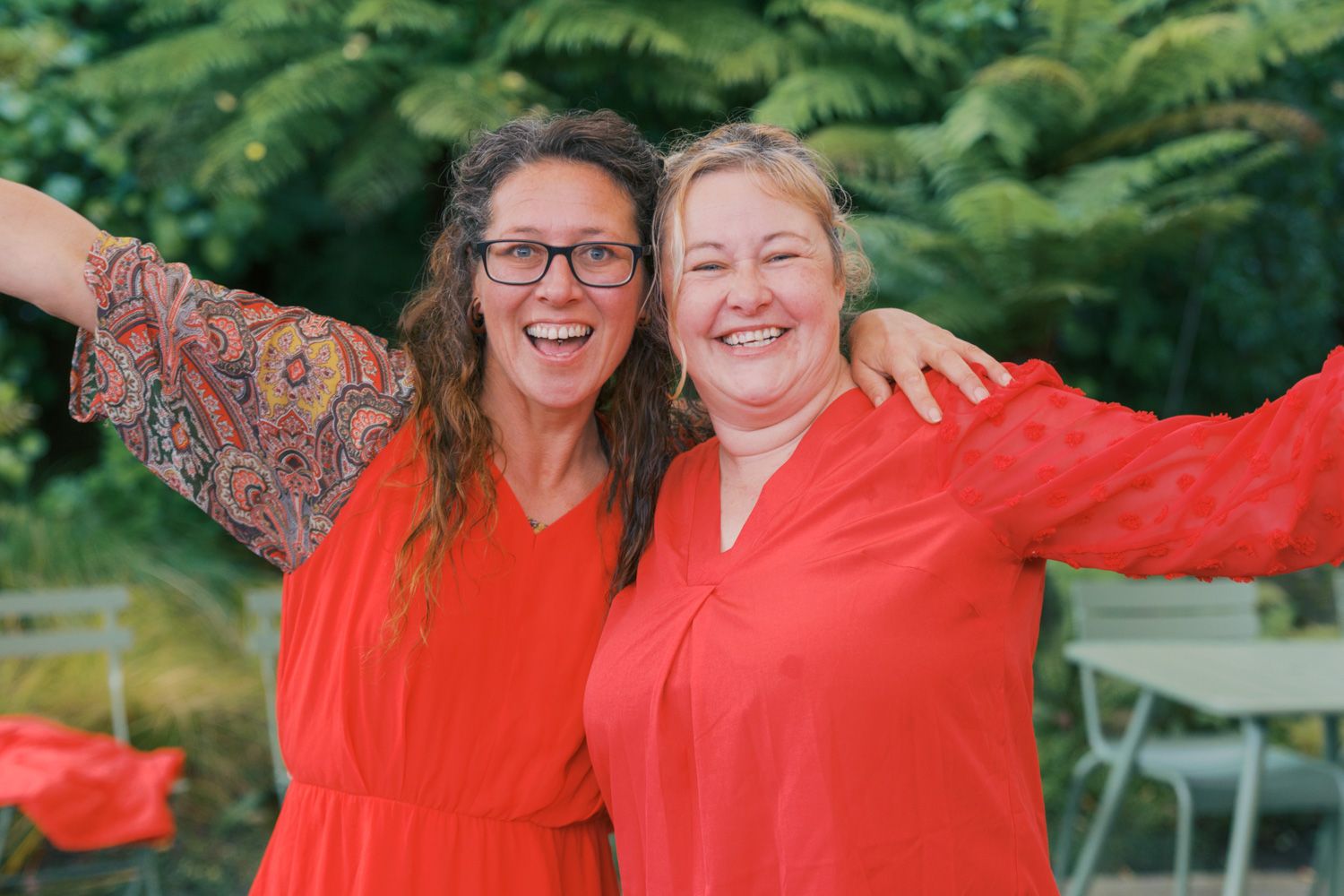 Two smiling women in matching red outfits embracing outdoors with green trees in the background.