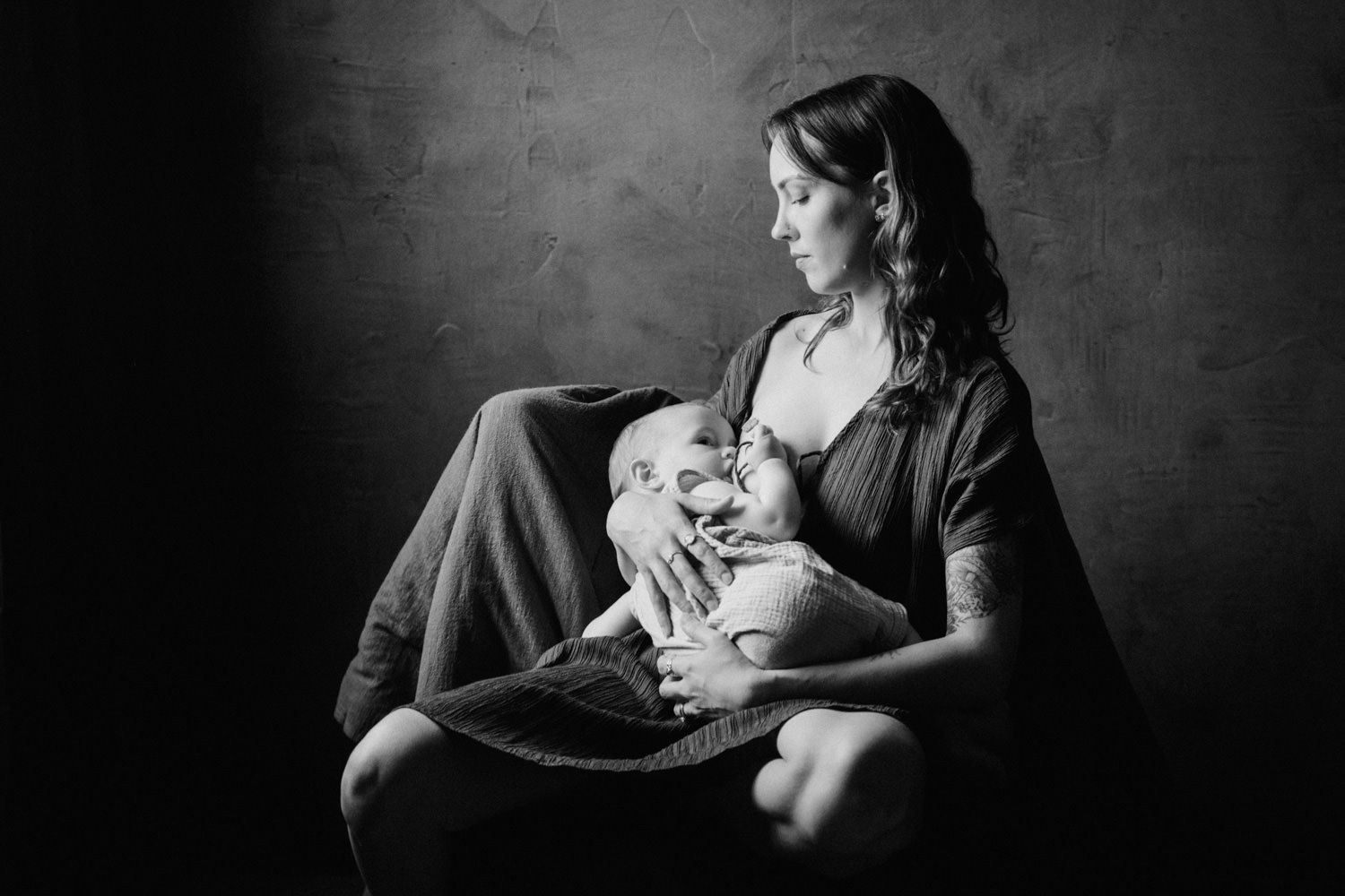 Black and white portrait of a mother breastfeeding her newborn baby while seated, captured in soft studio lighting.