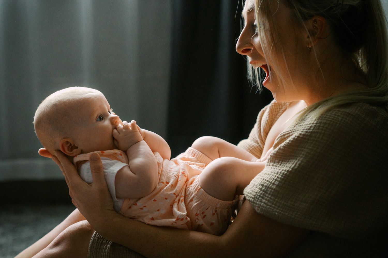 A smiling young mother holds her newborn baby in pink outfit in a softly lit room.