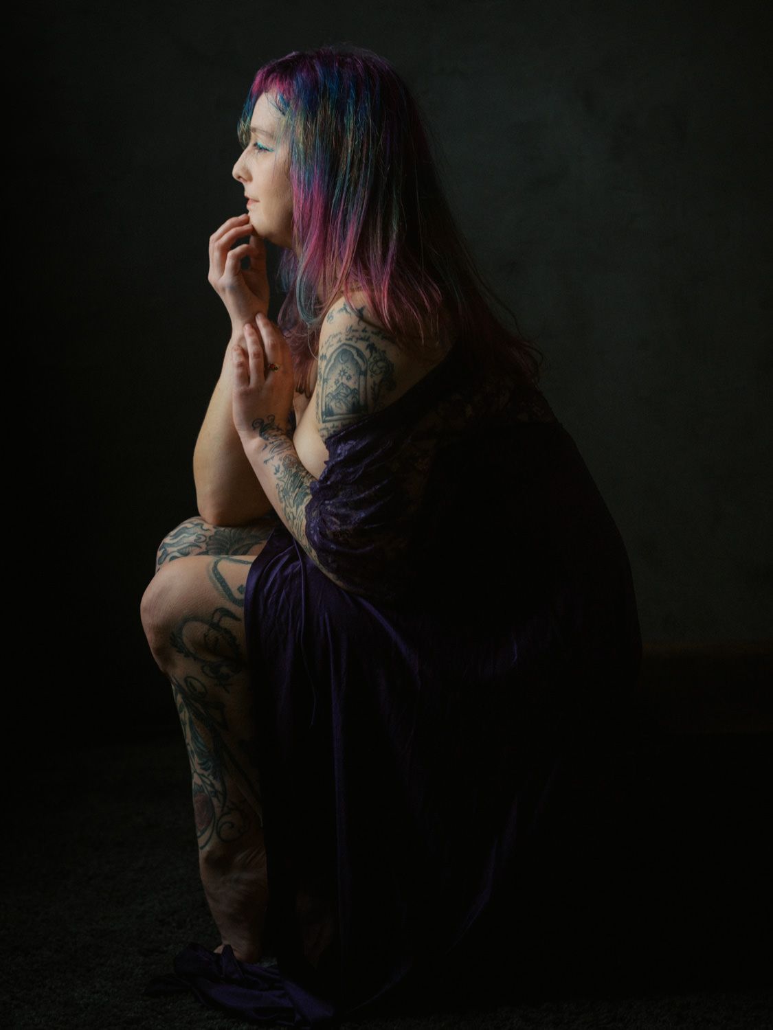 Woman with colorful pink-blue hair and tattoos sits pensively in a dark studio, wearing a purple dress.
