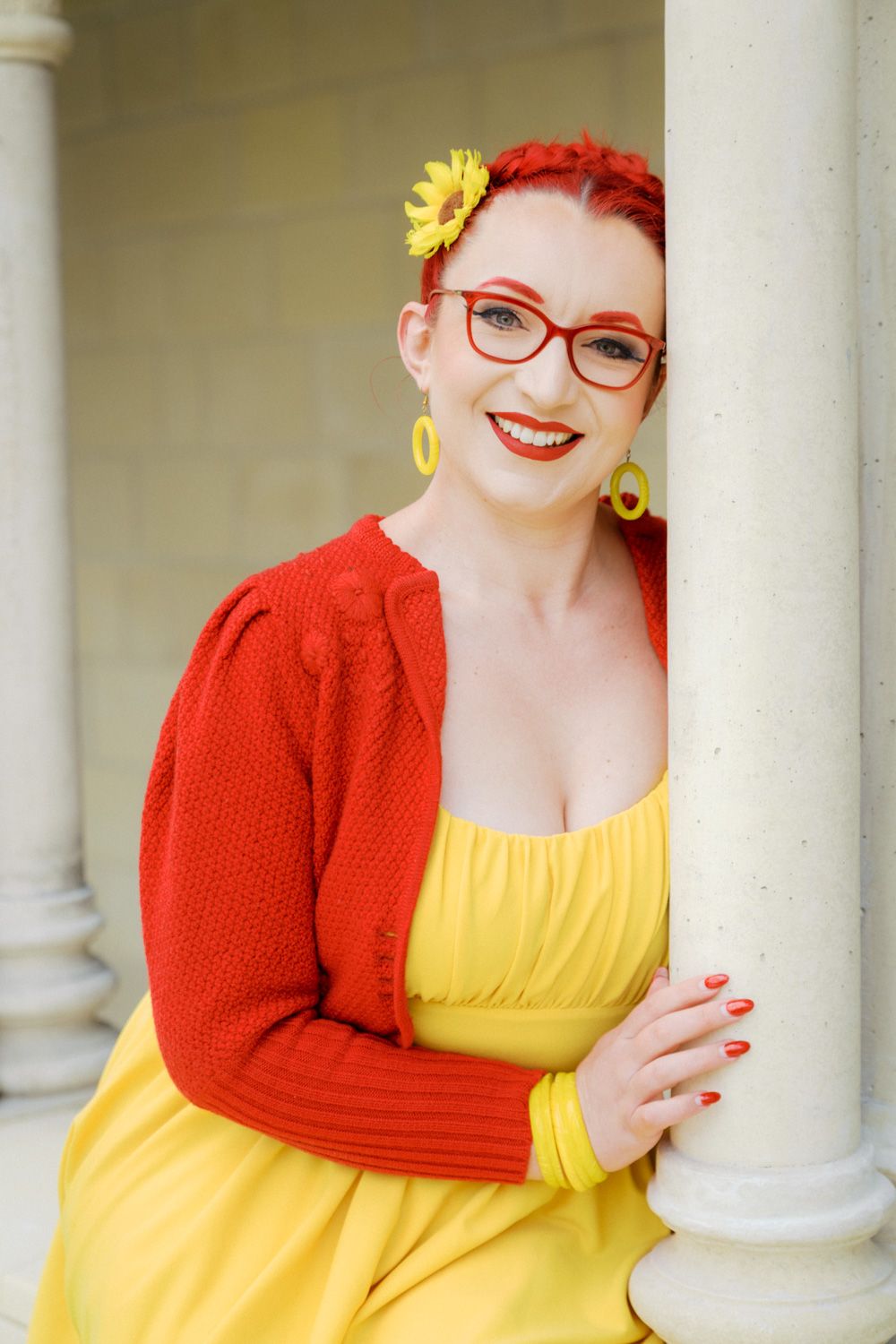 Smiling redhead woman in yellow dress and red cardigan, wearing red glasses and yellow flower hair accessory, leaning on white column.
