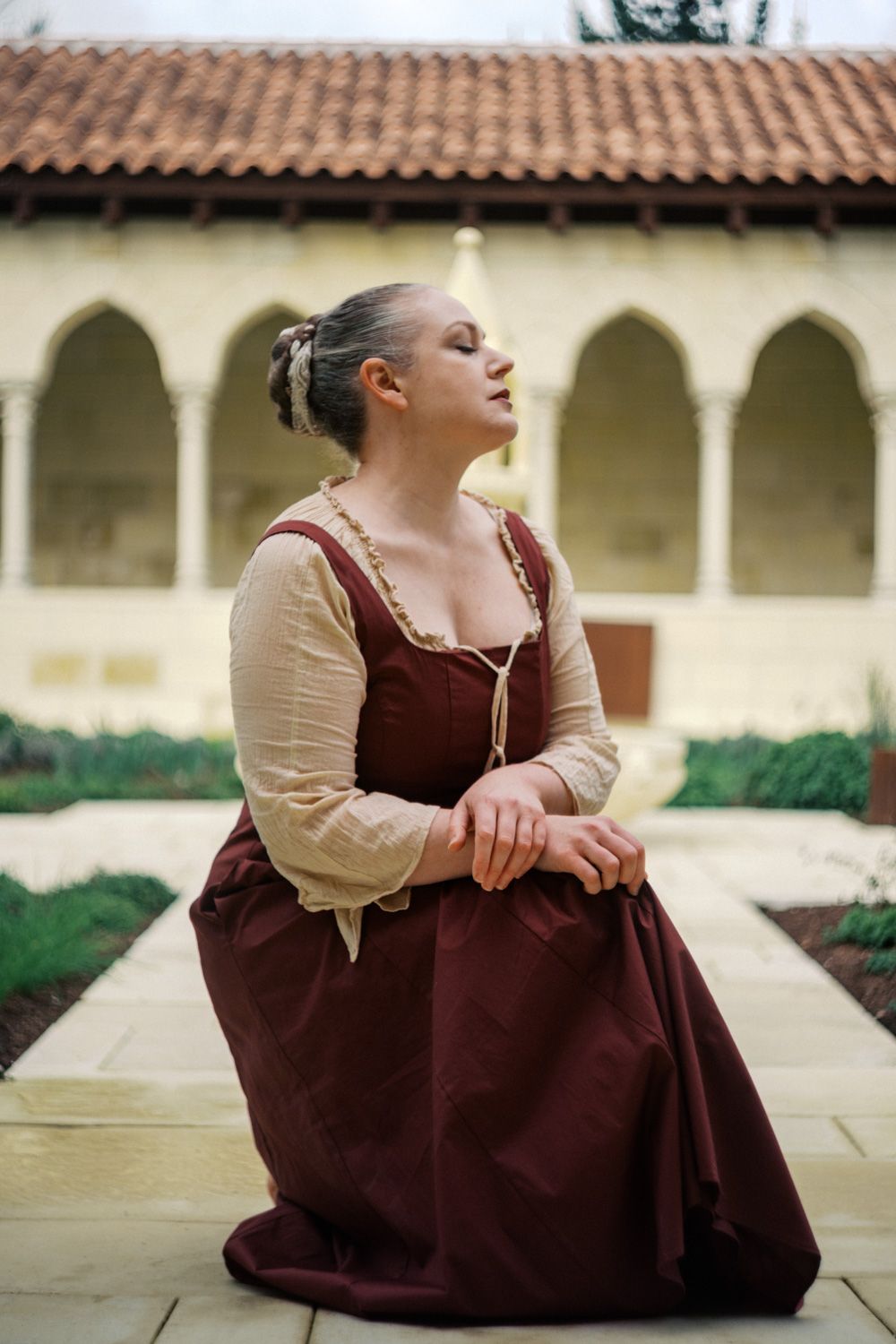 Woman in medieval costume kneeling in historic courtyard with arched colonnade and terracotta roof tiles.