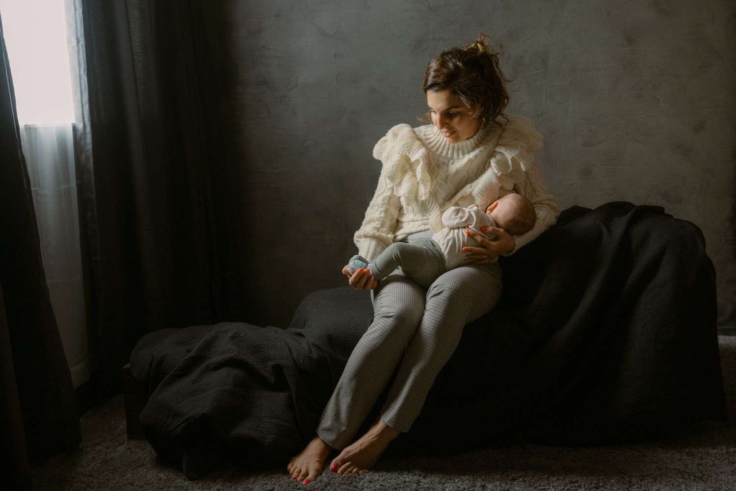 A young mother in a white knit sweater sits on a bean bag chair, gently cradling her newborn baby indoors.