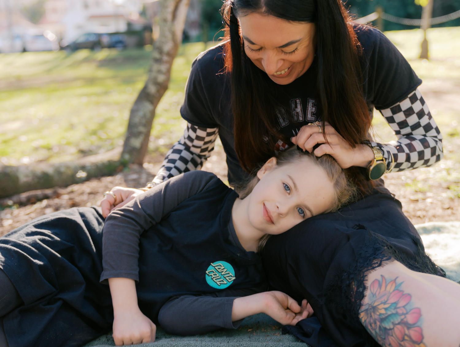Woman gently styling a child's hair as the child rests on a rock in a sunny park.