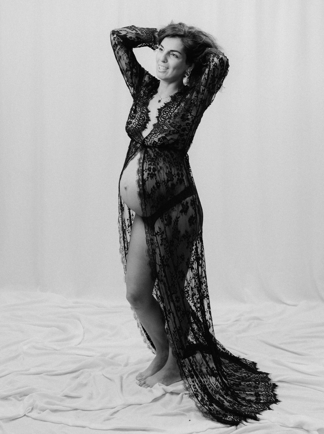 Pregnant woman in sheer black lace gown poses elegantly in black and white maternity photography session.